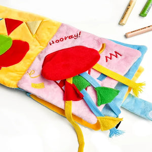 A colorful baby sensory activity book or soft fabric book spread open on a white background, showing textured panels in yellow, pink, blue, red, green, and white with embroidered text and ribbon tassels. A few crayons are partially visible at the top right.