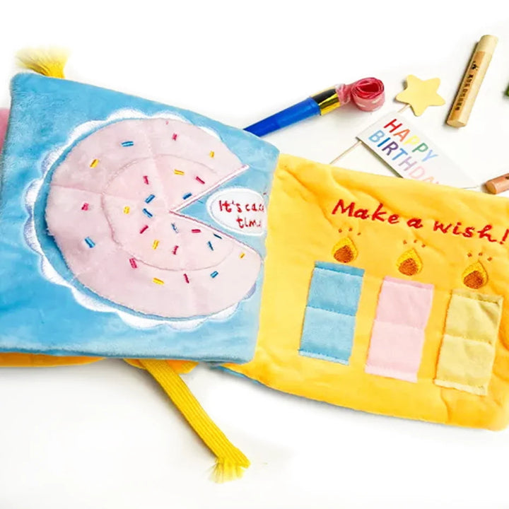 A plush fabric baby book with colorful panels, featuring a pink donut with sprinkles on a blue cover and a yellow page that says 'Make a wish!' with three colored squares. Party-themed props like a star, rose, and birthday candle are arranged around it on a white background.